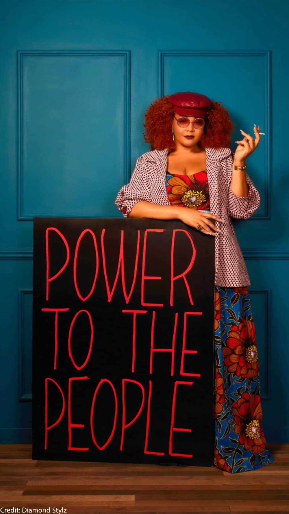 Diamond Stylz holds a cigarette as she leans on a sign that reads " POWER TO THE PEOPLE".