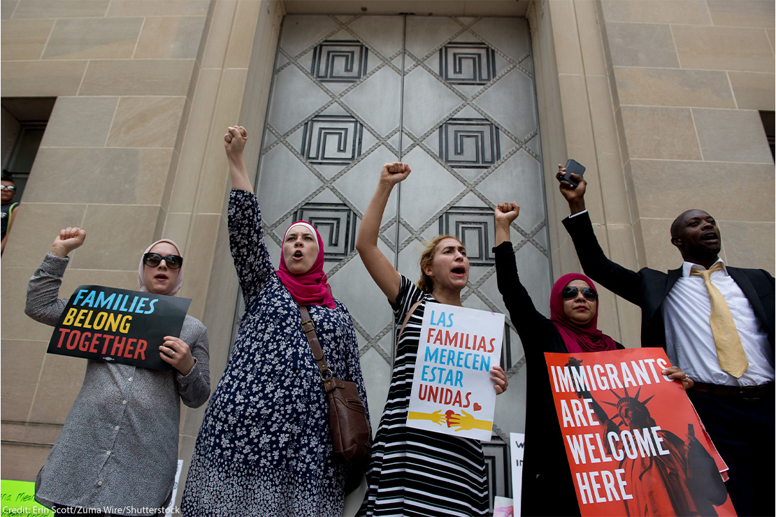 Protesters with closed fists raised holding signs reading "FAMILIES BELONG TOGETHER", LAS FAMILIAS MERECEN ESTAR UNIDAS" and IMMIGRANTS ARE WELCOME HERE" ACLU sign.