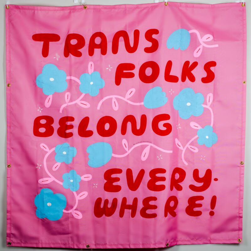 A four color (pink, white, red, blue) illustrated acrylic panel featuring interwoven flowers witht he message "trans folk belong everywhere".