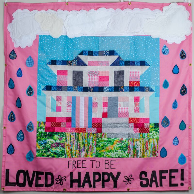 Pink panel with quilted block depicting a pink and blue hosue with a trans flag out front on the porch with green grass and a blue sky. Above the blcok there are clouds and there are blue raindrops on either side. Black text at the bottom reads "Free to Be: Loved, Happy, Safe"