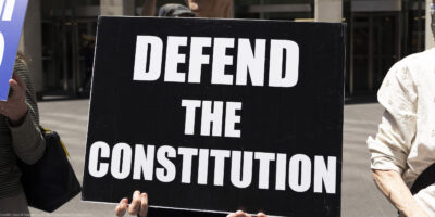 A demonstrator holds a sign (with white letters on a black background) that reads "DEFEND THE CONSTITUTION".