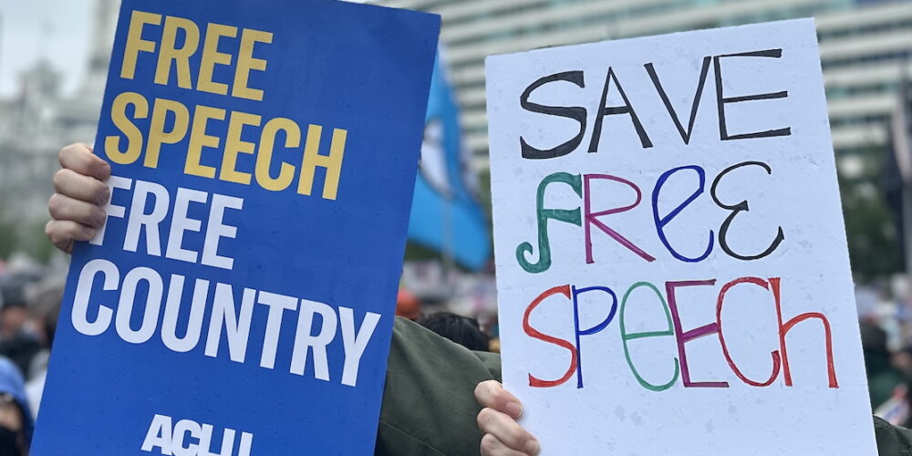 One protest sign reads: "Free Speech, Free Country." The other reads: "Save Free Speech"