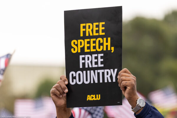 A demonstrator (with only their hands visible) holds up a sign that reads " FREE SPEECH, FREE COUNTRY - ACLU".