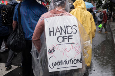A person wearing a clear poncho displays a sign on their back that reads "HANDS OFF THE CONSTITUTION" while walking in the rain as they take part with thousands of other New Yorkers take part in the NO KINGS national day of action.