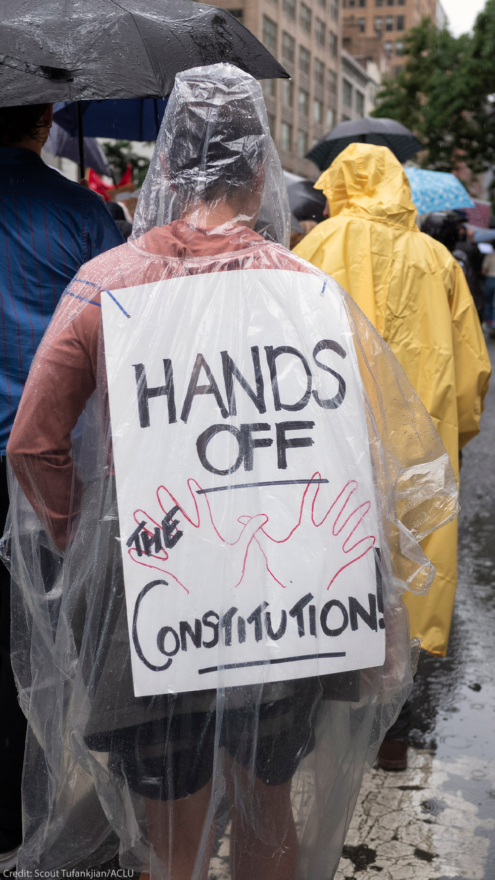 A person wearing a clear poncho displays a sign on their back that reads "HANDS OFF THE CONSTITUTION" while walking in the rain as they take part with thousands of other New Yorkers take part in the NO KINGS national day of action.