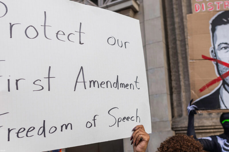A person holds a large handwritten sign reading “Protect our First Amendment Freedom of Speech” during a protest outside the El Capitan Entertainment Centre, where Jimmy Kimmel Live! is filmed. Behind them, another protester in a skeleton costume holds a cardboard sign depicting a crossed-out image of Jimmy Kimmel. The demonstration is in response to ABC’s decision to pull Kimmel off the air following his remarks about Charlie Kirk.
