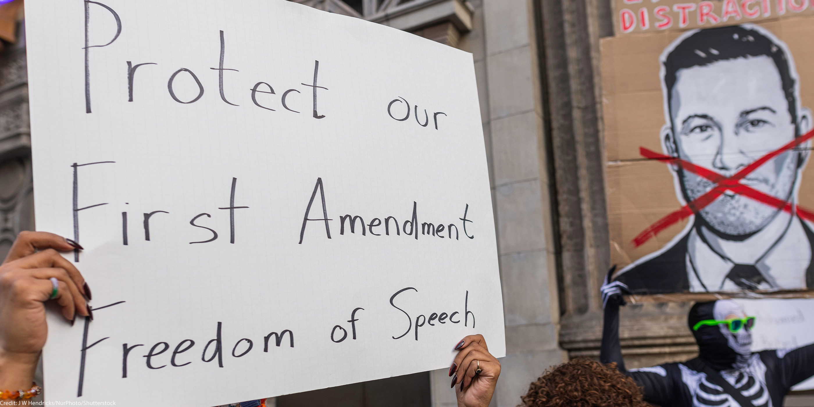 A person holds a large handwritten sign reading “Protect our First Amendment Freedom of Speech” during a protest outside the El Capitan Entertainment Centre, where Jimmy Kimmel Live! is filmed. Behind them, another protester in a skeleton costume holds a cardboard sign depicting a crossed-out image of Jimmy Kimmel. The demonstration is in response to ABC’s decision to pull Kimmel off the air following his remarks about Charlie Kirk.