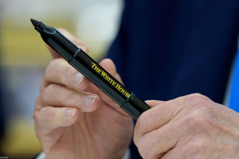 A close-up of President Trump’s hands holding a black marker pen inscribed with “The White House” in gold lettering, moments after signing executive orders in the Oval Office.