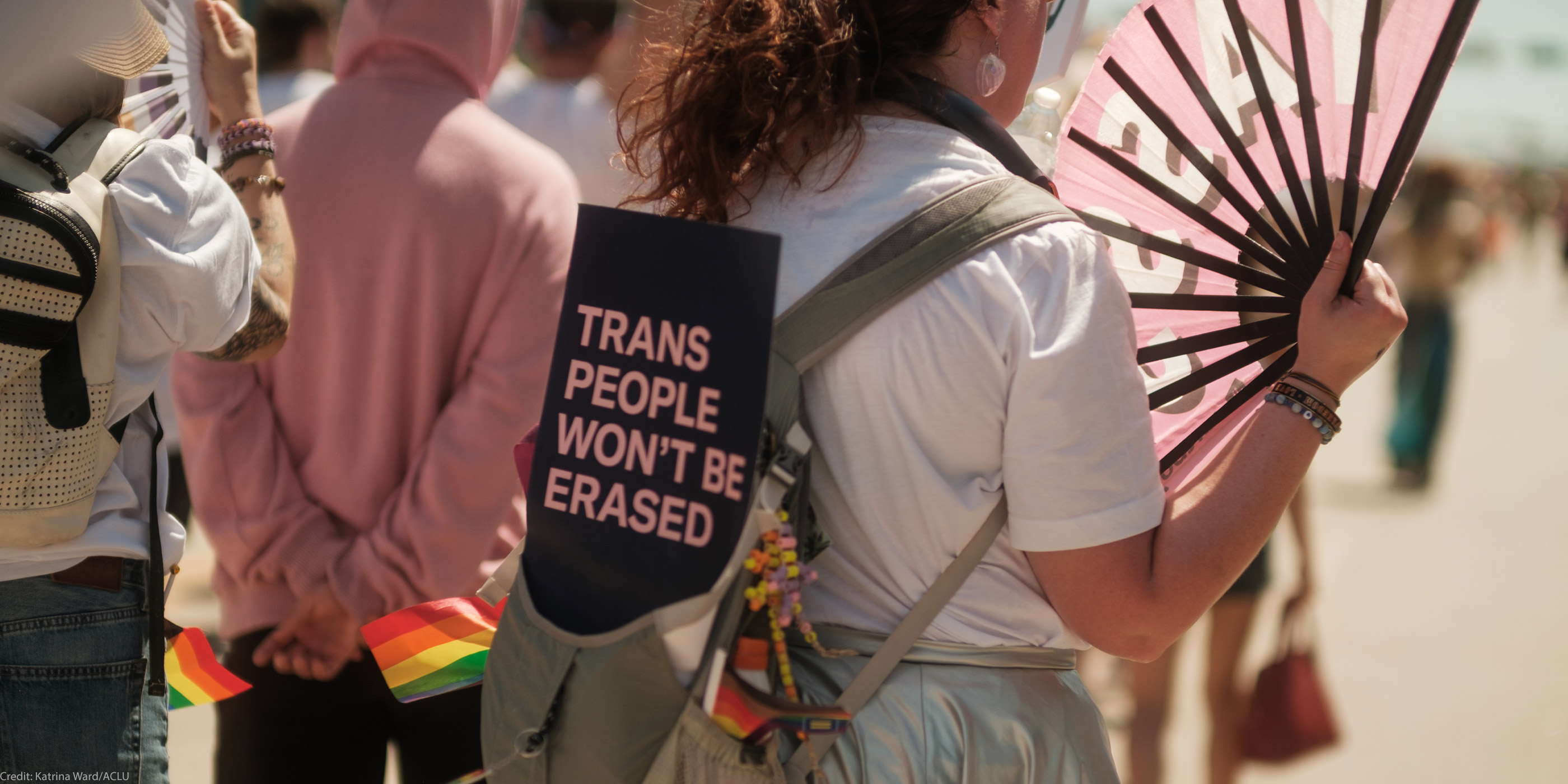 A person at an outdoor LGBTQ+ pride event carries a sign on their backpack that reads “Trans People Won’t Be Erased.” Small rainbow flags and a decorative bouquet are tucked into the backpack. The person holds a large pink fan, and other participants walk ahead of them in the crowd.