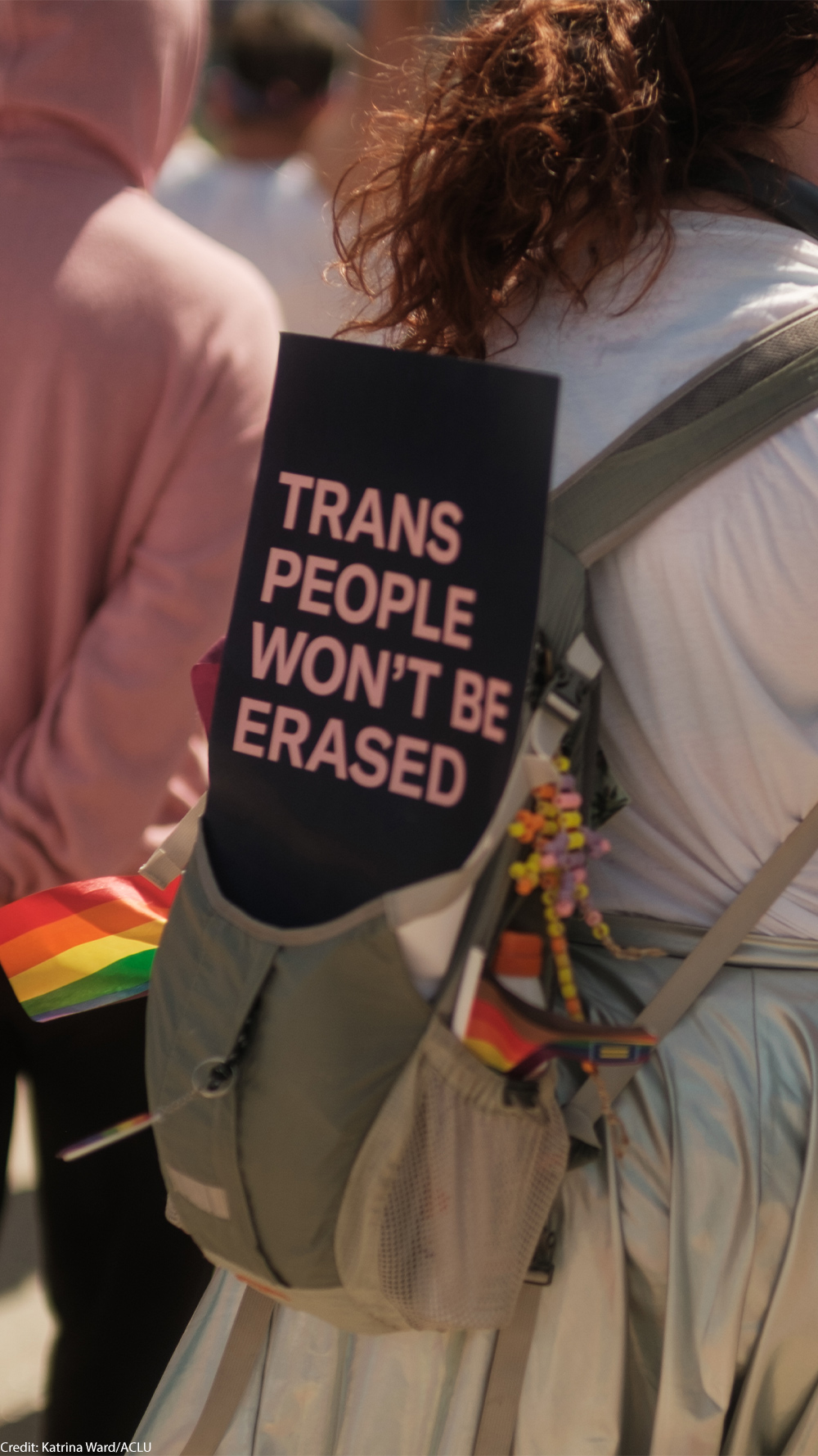 A person at an outdoor LGBTQ+ pride event carries a sign on their backpack that reads “Trans People Won’t Be Erased.” Small rainbow flags and a decorative bouquet are tucked into the backpack. The person holds a large pink fan, and other participants walk ahead of them in the crowd.