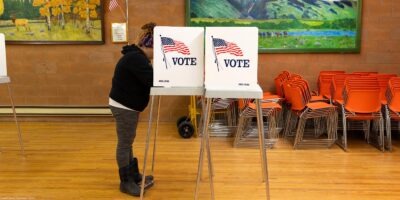 A person stands at a voting booth marked with an American flag and the word “VOTE” inside a community polling place. The room has wooden floors, orange stacked chairs, and colorful landscape paintings on the walls, with a small U.S. flag displayed nearby.