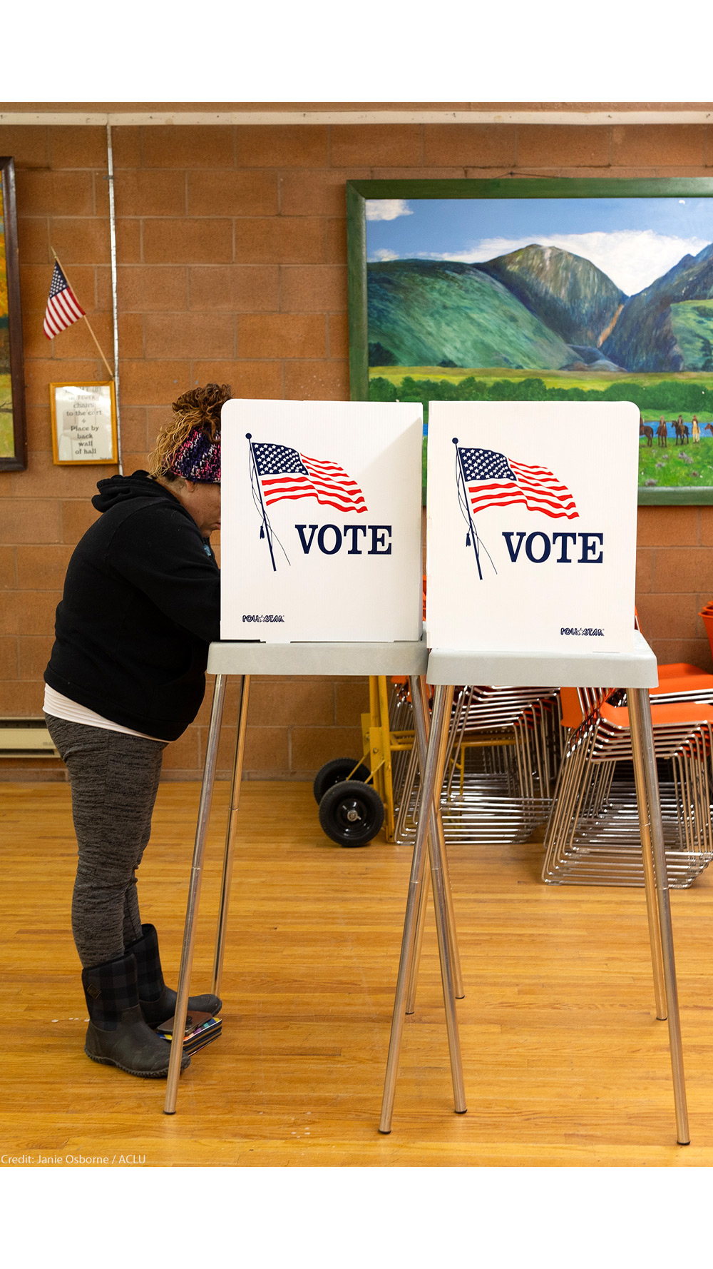 A person stands at a voting booth marked with an American flag and the word “VOTE” inside a community polling place. The room has wooden floors, orange stacked chairs, and colorful landscape paintings on the walls, with a small U.S. flag displayed nearby.