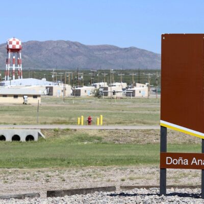 A large brown U.S. Army sign reading “Fort Bliss – Doña Ana Training Complex” stands in the foreground of a wide, open landscape. Behind it are low beige military buildings, utility poles, and a tall red-and-white checkered water tower, with mountains rising in the distance under a clear blue sky.