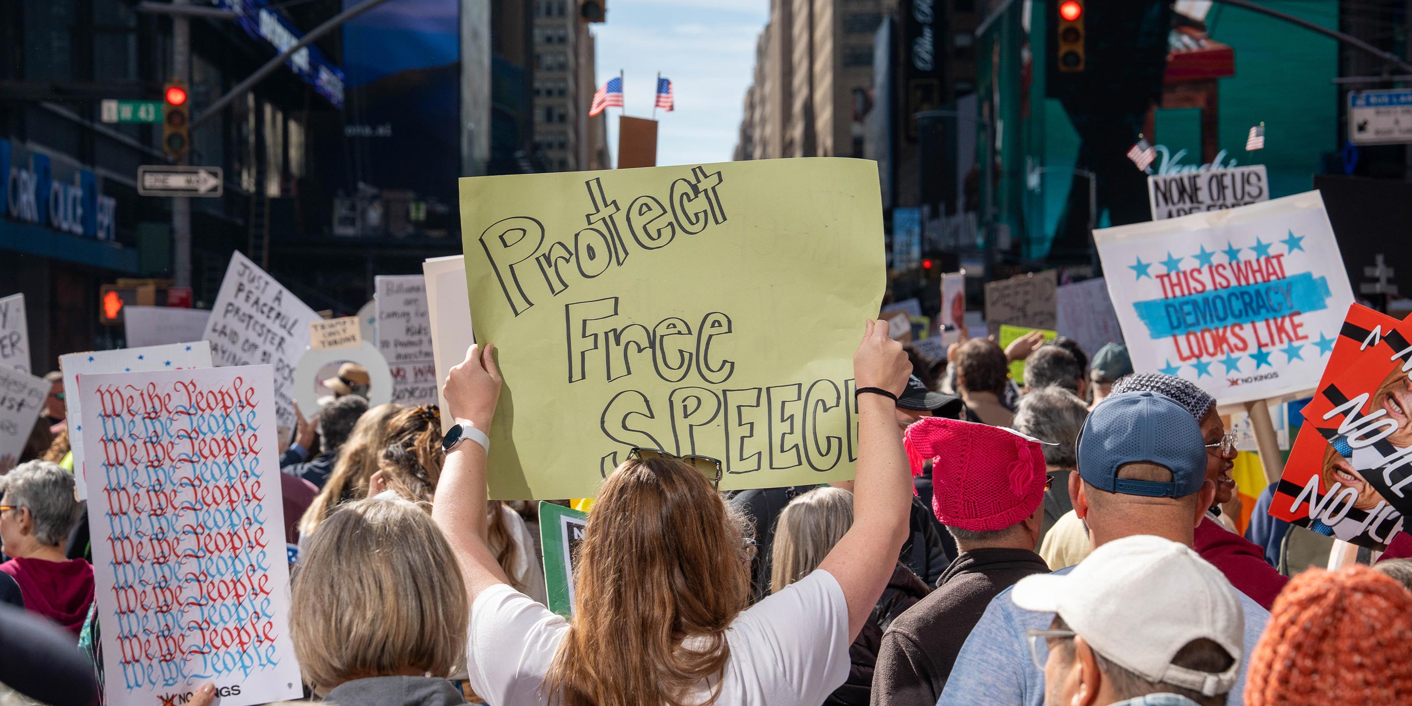 A large crowd of demonstrators fills a city street, with one person in the foreground holding a bright yellow sign that reads “Protect Free Speech.” Other protest signs are visible throughout the crowd, including messages such as “We the People” and “This Is What Democracy Looks Like.”