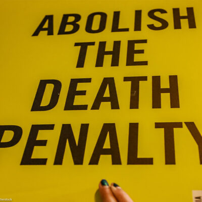 Yellow protest sign with bold black text reading “Abolish the Death Penalty,” partially held by a person at the bottom edge.