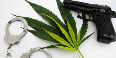 Handcuffs and a handgun next to cannabis leaves on a white background.