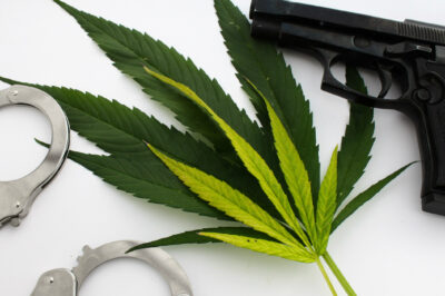 Handcuffs and a handgun next to cannabis leaves on a white background.