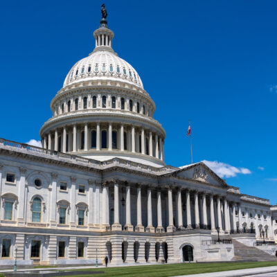 Wide daytime view of the U.S. Capitol building in Washington, D.C., with the white dome and columns under a bright blue sky.
