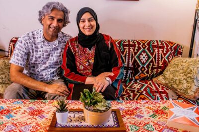 A smiling Dr. Badar Khan Suri, wearing a light, patterned short-sleeve button-down shirt and his wife, wearing a black headscarf with a red-and-black embroidered top, sit on a patterned sofa in their cozy living room, which has a colorful tablecloth and small potted plants on the coffee table in front of them.