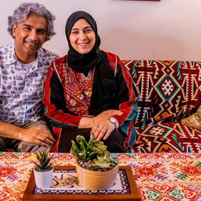 A smiling Dr. Badar Khan Suri, wearing a light, patterned short-sleeve button-down shirt and his wife, wearing a black headscarf with a red-and-black embroidered top, sit on a patterned sofa in their cozy living room, which has a colorful tablecloth and small potted plants on the coffee table in front of them.