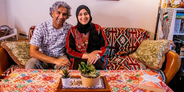 A smiling Dr. Badar Khan Suri, wearing a light, patterned short-sleeve button-down shirt and his wife, wearing a black headscarf with a red-and-black embroidered top, sit on a patterned sofa in their cozy living room, which has a colorful tablecloth and small potted plants on the coffee table in front of them.
