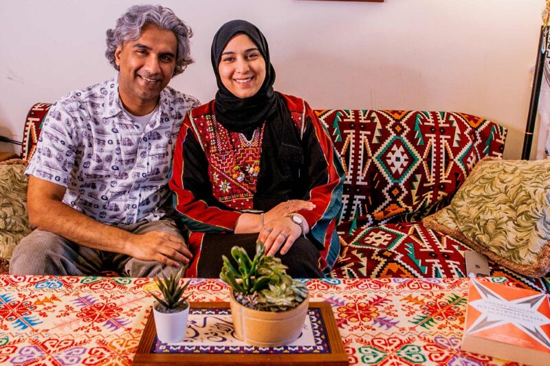 A smiling Dr. Badar Khan Suri, wearing a light, patterned short-sleeve button-down shirt and his wife, wearing a black headscarf with a red-and-black embroidered top, sit on a patterned sofa in their cozy living room, which has a colorful tablecloth and small potted plants on the coffee table in front of them.