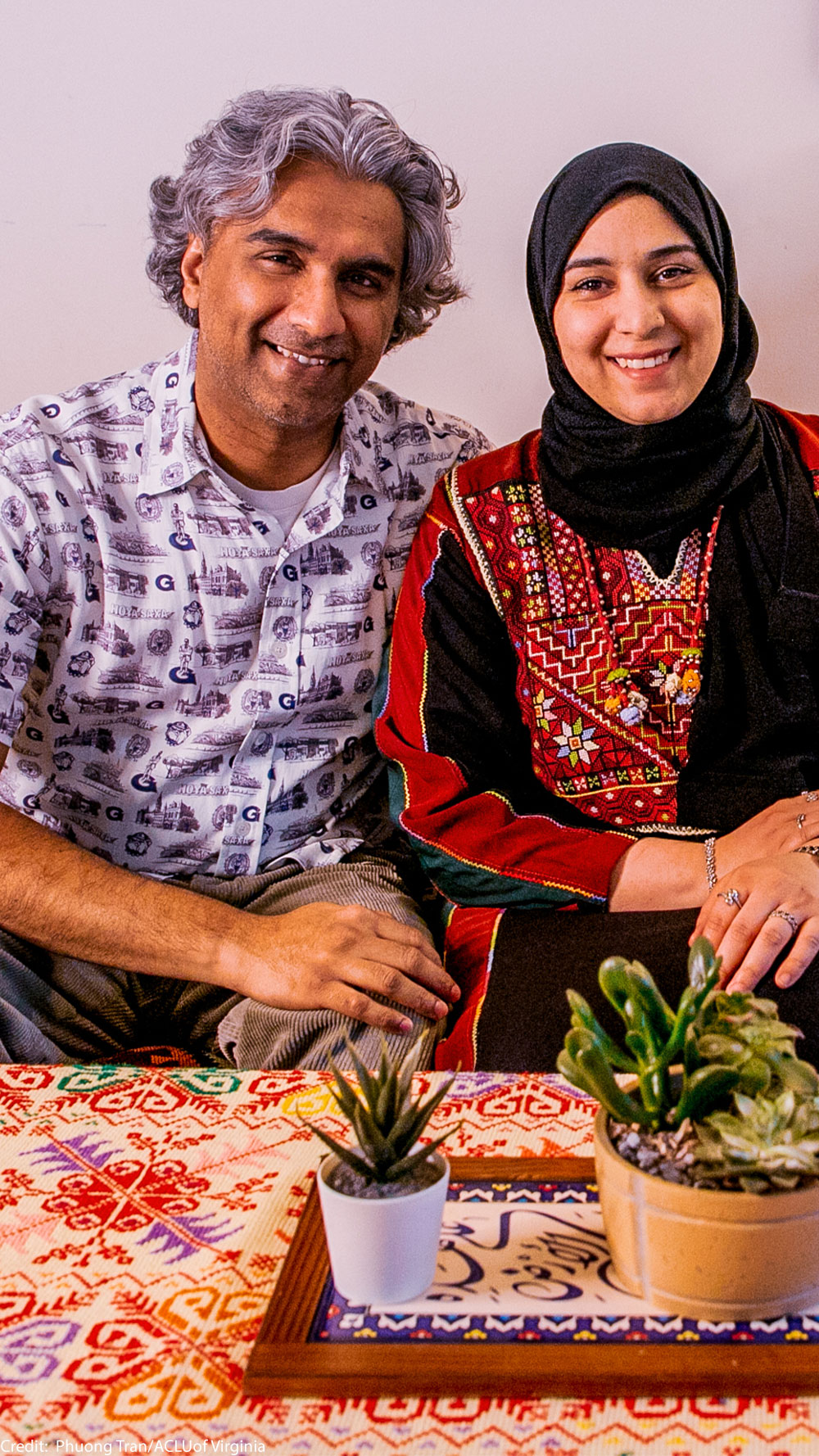 A smiling Dr. Badar Khan Suri, wearing a light, patterned short-sleeve button-down shirt and his wife, wearing a black headscarf with a red-and-black embroidered top, sit on a patterned sofa in their cozy living room, which has a colorful tablecloth and small potted plants on the coffee table in front of them.