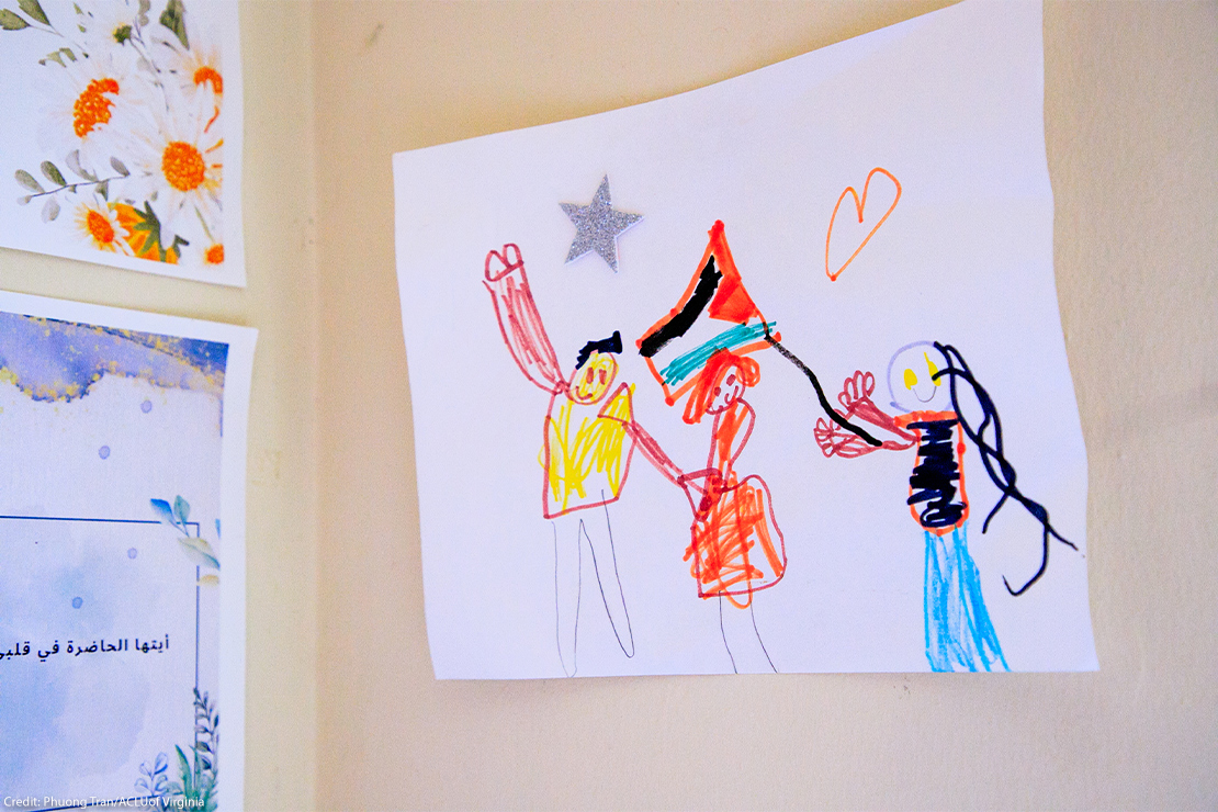 Child’s crayon drawing taped to a wall showing three figures holding hands, one waving a Palestinian flag, with a star and heart drawn above.