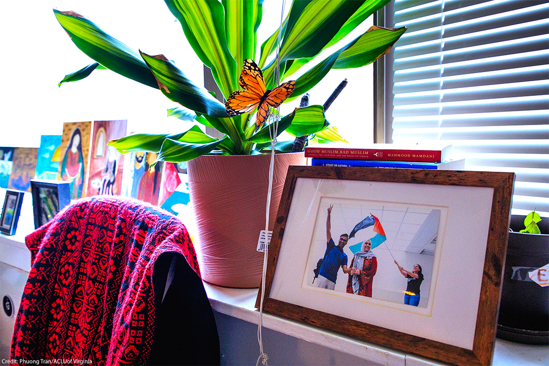 A windowsill with a potted green plant and an orange butterfly decoration beside a framed photo of three smiling people holding a Palestinian flag (with books and colorful artwork in the background). Within the frame, Dr. Badar Khan Suri is raising a peace sign, a woman in a headscarf is standing in the center, and another person is holding the flagpole.