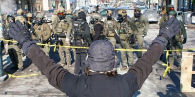 A person (with their back to the camera) in a winter hat and gloves stands with both arms raised during an anti-ICE demonstration, facing a line of police officers in tactical gear behind yellow tape on a Minneapolis street.