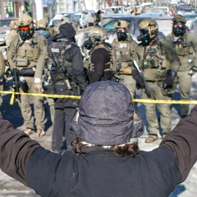 A person (with their back to the camera) in a winter hat and gloves stands with both arms raised during an anti-ICE demonstration, facing a line of police officers in tactical gear behind yellow tape on a Minneapolis street.