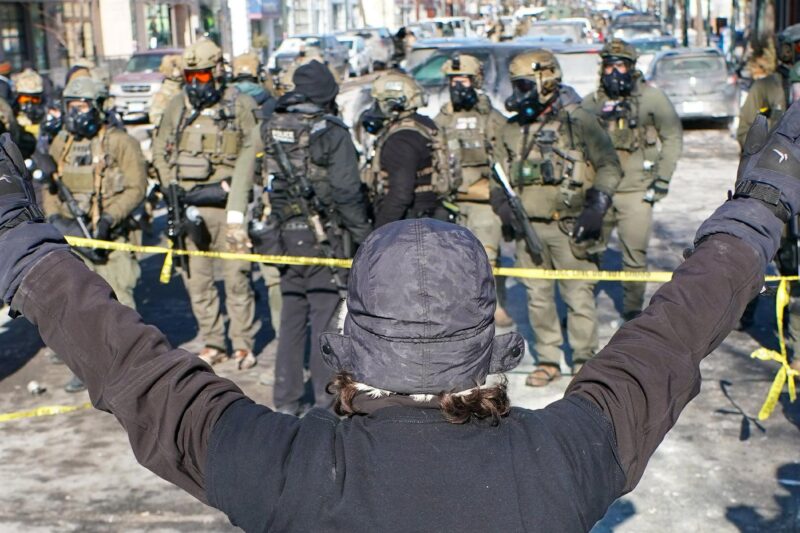 A person (with their back to the camera) in a winter hat and gloves stands with both arms raised during an anti-ICE demonstration, facing a line of police officers in tactical gear behind yellow tape on a Minneapolis street.