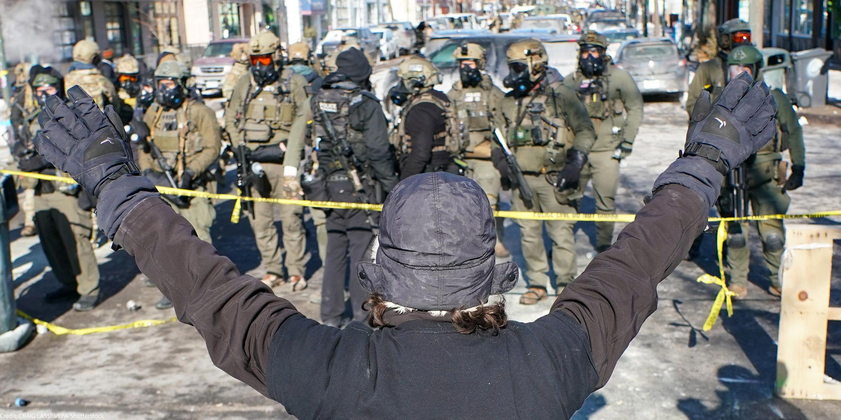 A person (with their back to the camera) in a winter hat and gloves stands with both arms raised during an anti-ICE demonstration, facing a line of police officers in tactical gear behind yellow tape on a Minneapolis street.