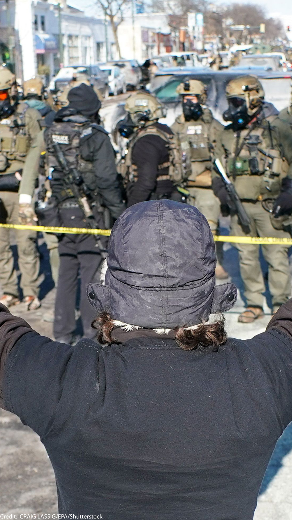 A person (with their back to the camera) in a winter hat and gloves stands with both arms raised during an anti-ICE demonstration, facing a line of police officers in tactical gear behind yellow tape on a Minneapolis street.