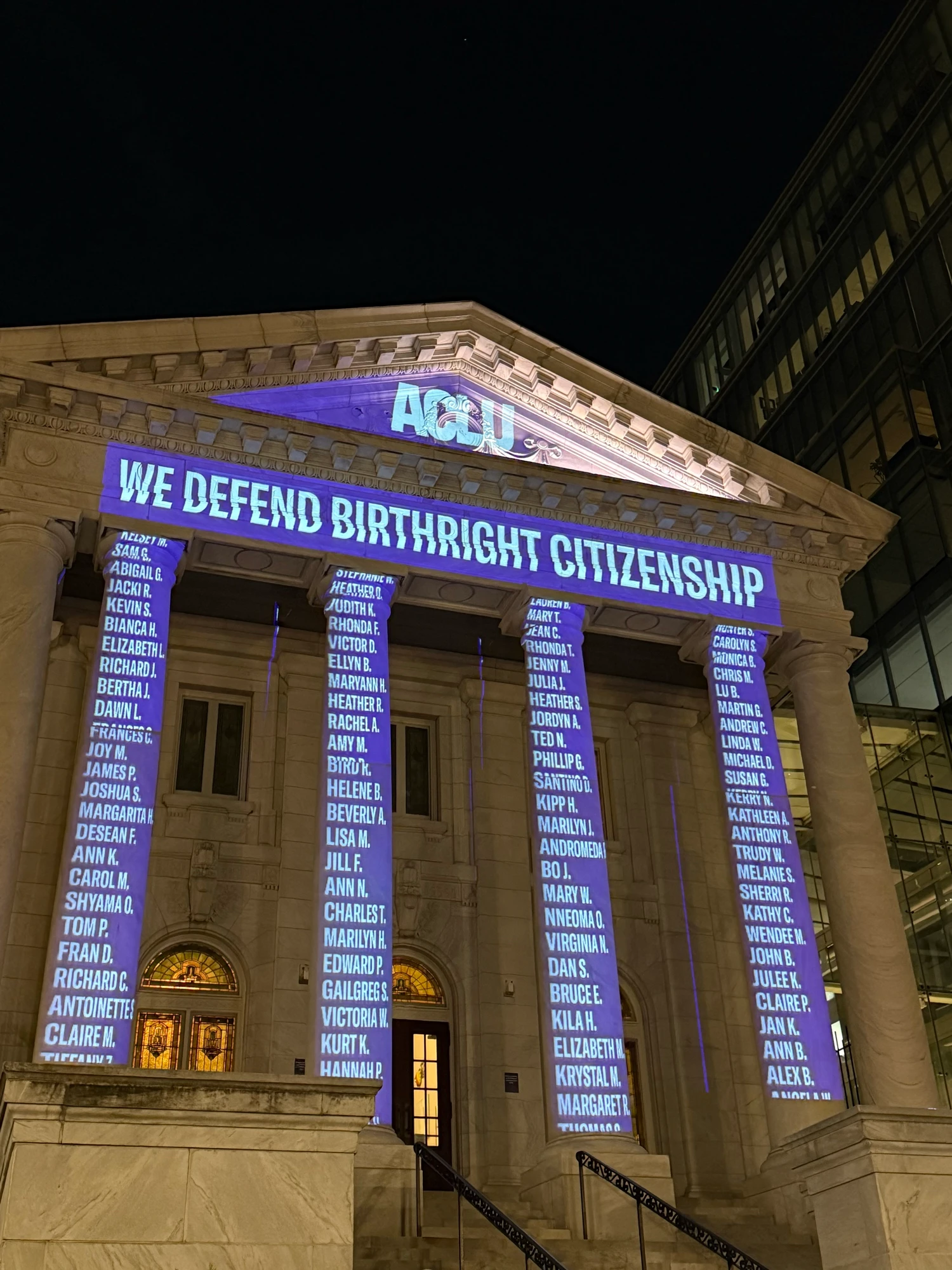 Light projection on building with columns that says We Defend Birthright Citizenship and has hundreds of names.
