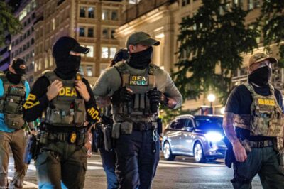 Four masked DEA police officers wearing tactical vests labeled “DEA POLICE” walk across a downtown intersection at night, with tall office buildings, streetlights, traffic signals, and cars with headlights behind them.
