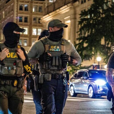Four masked DEA police officers wearing tactical vests labeled “DEA POLICE” walk across a downtown intersection at night, with tall office buildings, streetlights, traffic signals, and cars with headlights behind them.
