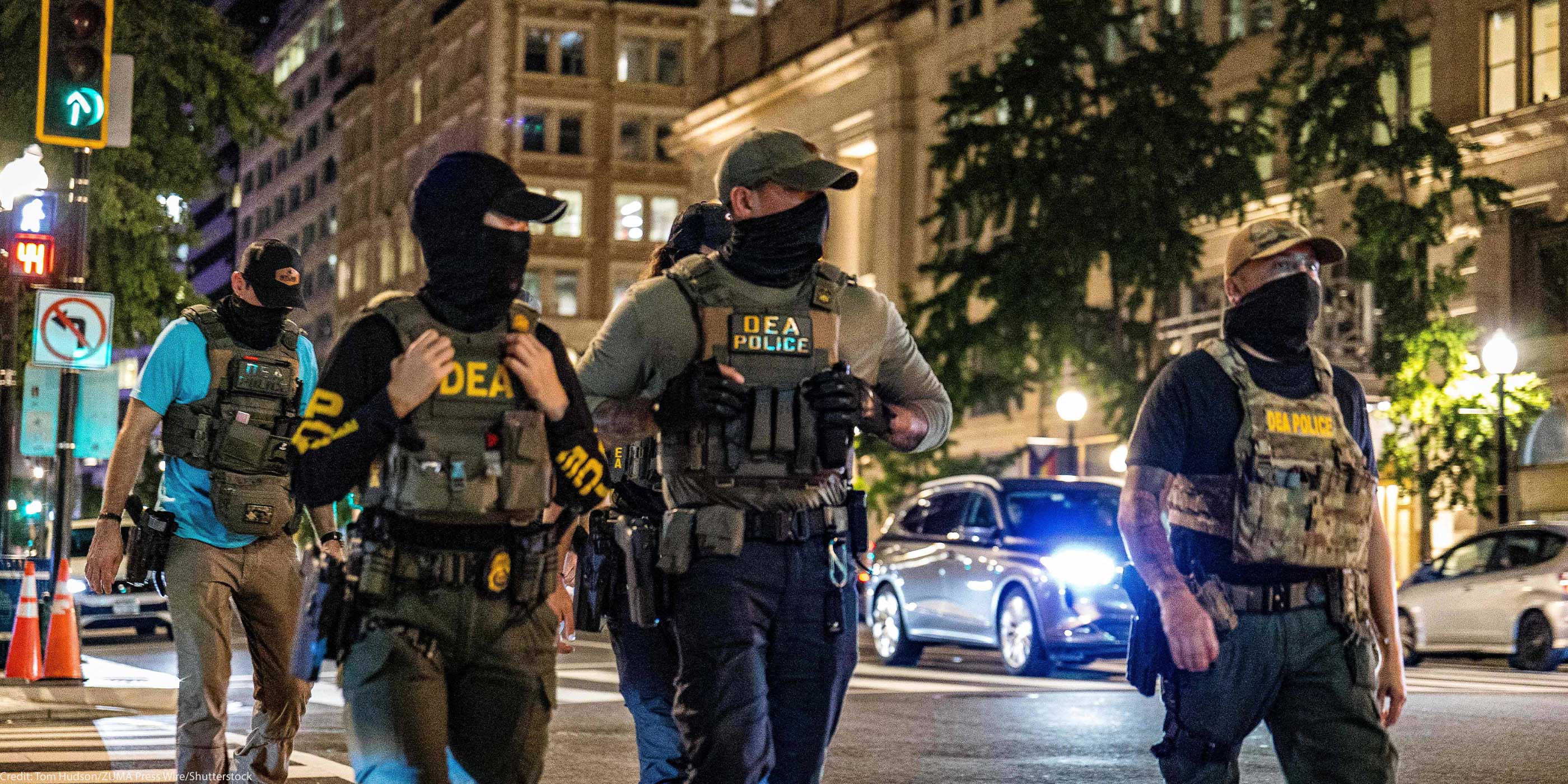 Four masked DEA police officers wearing tactical vests labeled “DEA POLICE” walk across a downtown intersection at night, with tall office buildings, streetlights, traffic signals, and cars with headlights behind them.