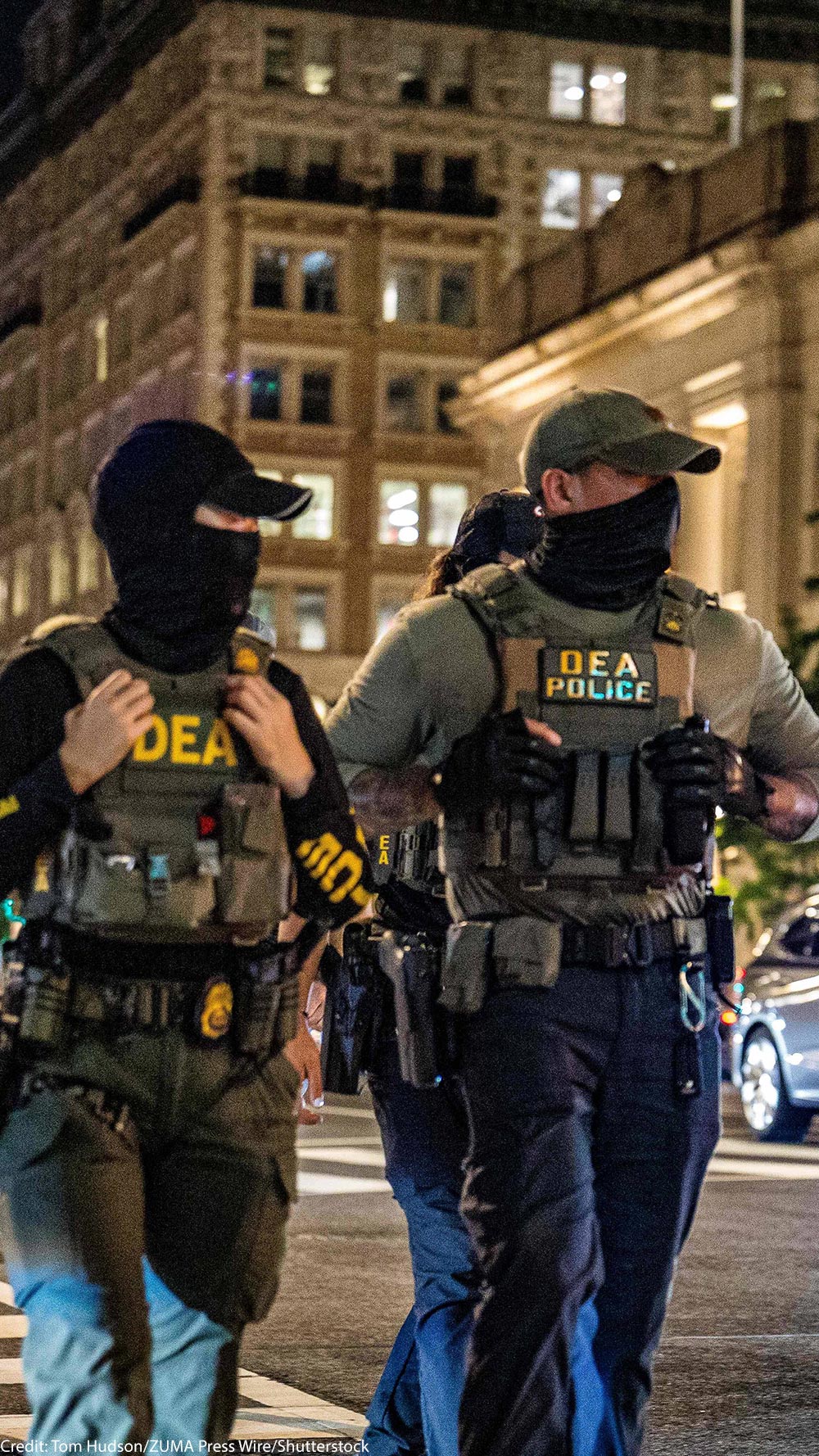 Two masked DEA police officers wearing tactical vests labeled “DEA POLICE” walk across a downtown intersection at night, with tall office buildings, streetlights, traffic signals, and cars with headlights behind them.