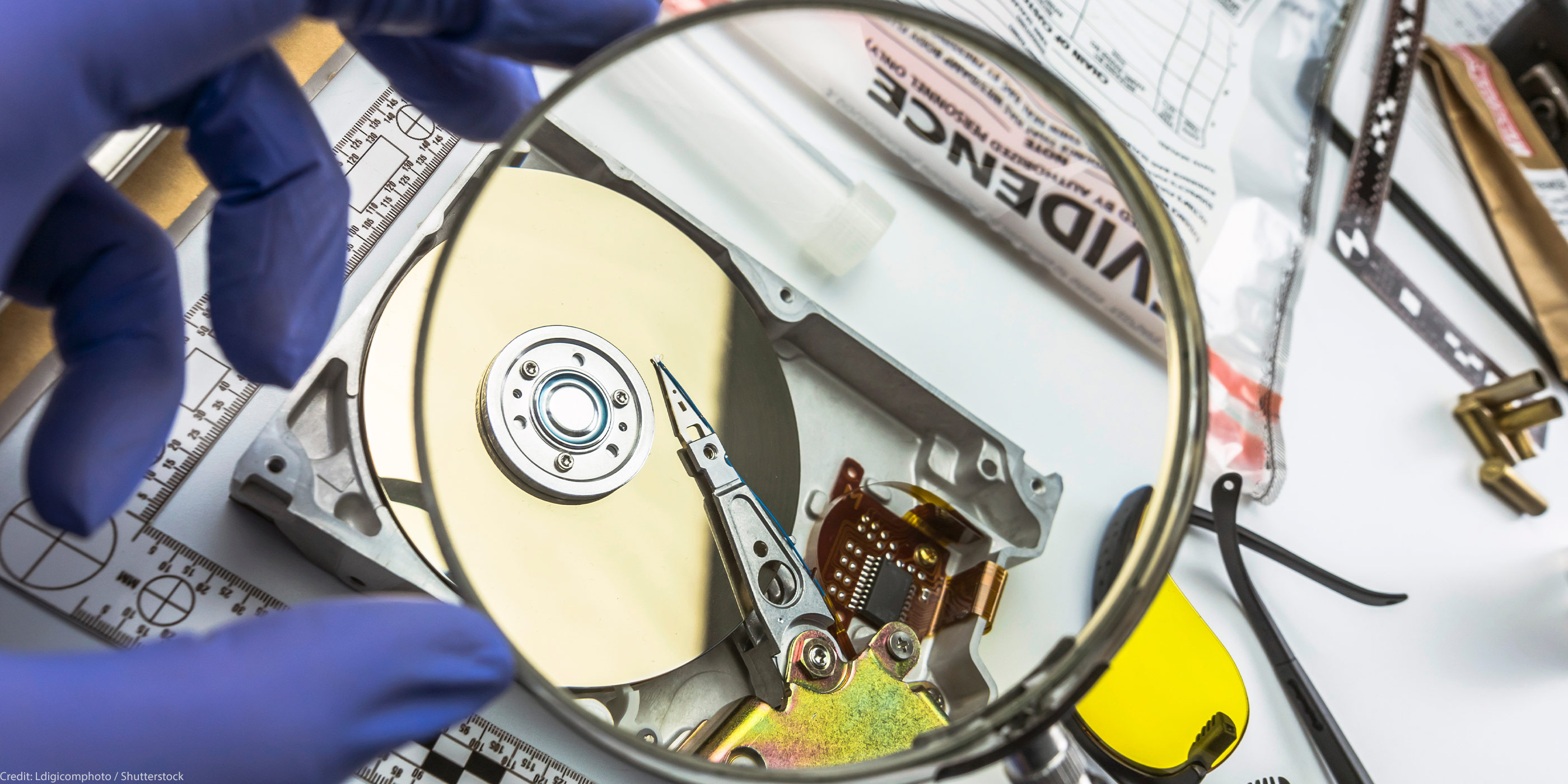Gloved hands hold a magnifying glass over an opened computer hard drive on a forensic work surface, with evidence bags, tools, and safety glasses nearby.