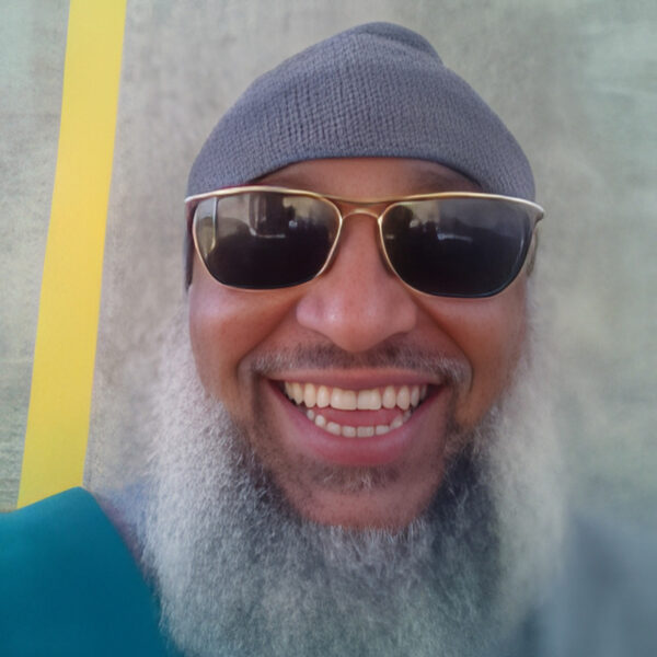 Close-up portrait of a smiling Tony Carruthers with a full gray beard, wearing dark sunglasses and a gray knit cap against a light background.
