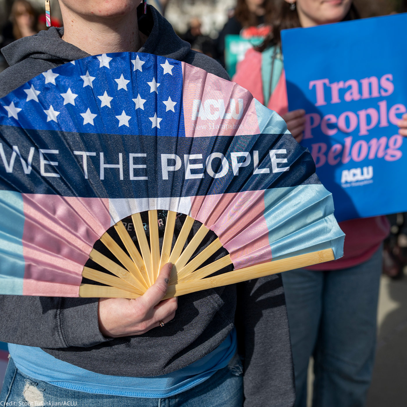 A protester in a gray hoodie holds an open fan in transgender flag colors that reads "WE THE PEOPLE," while others stand behind them with a blue sign reading "Trans People Belong" (ACLU).