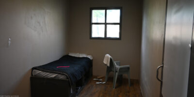 A small, sparsely furnished room with a single bed, a plastic chair with a towel draped over it, a barred window, and a pair of flip-flops on the wooden floor.