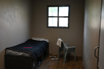 A small, sparsely furnished room with a single bed, a plastic chair with a towel draped over it, a barred window, and a pair of flip-flops on the wooden floor.