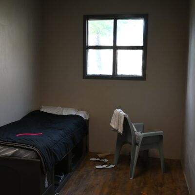 A small, sparsely furnished room with a single bed, a plastic chair with a towel draped over it, a barred window, and a pair of flip-flops on the wooden floor.