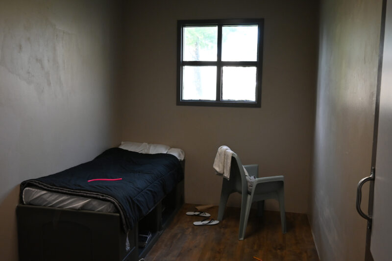 A small, sparsely furnished room with a single bed, a plastic chair with a towel draped over it, a barred window, and a pair of flip-flops on the wooden floor.
