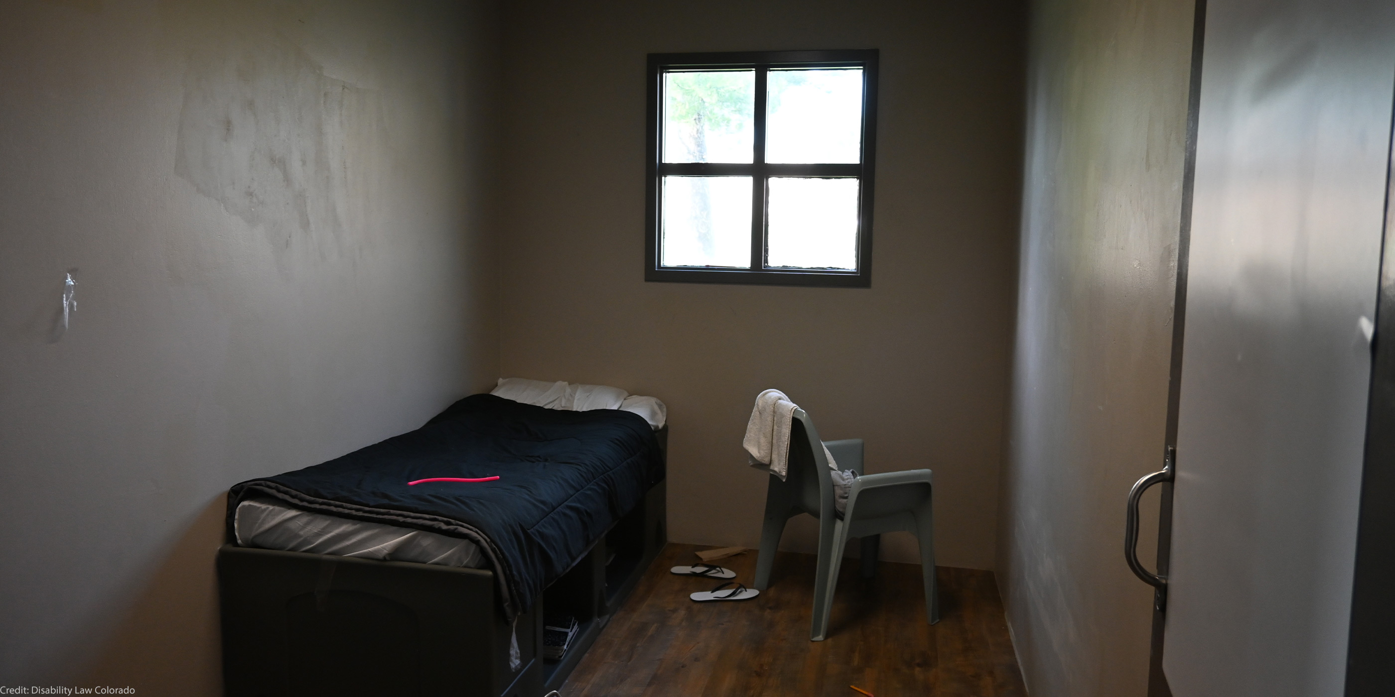 A small, sparsely furnished room with a single bed, a plastic chair with a towel draped over it, a barred window, and a pair of flip-flops on the wooden floor.