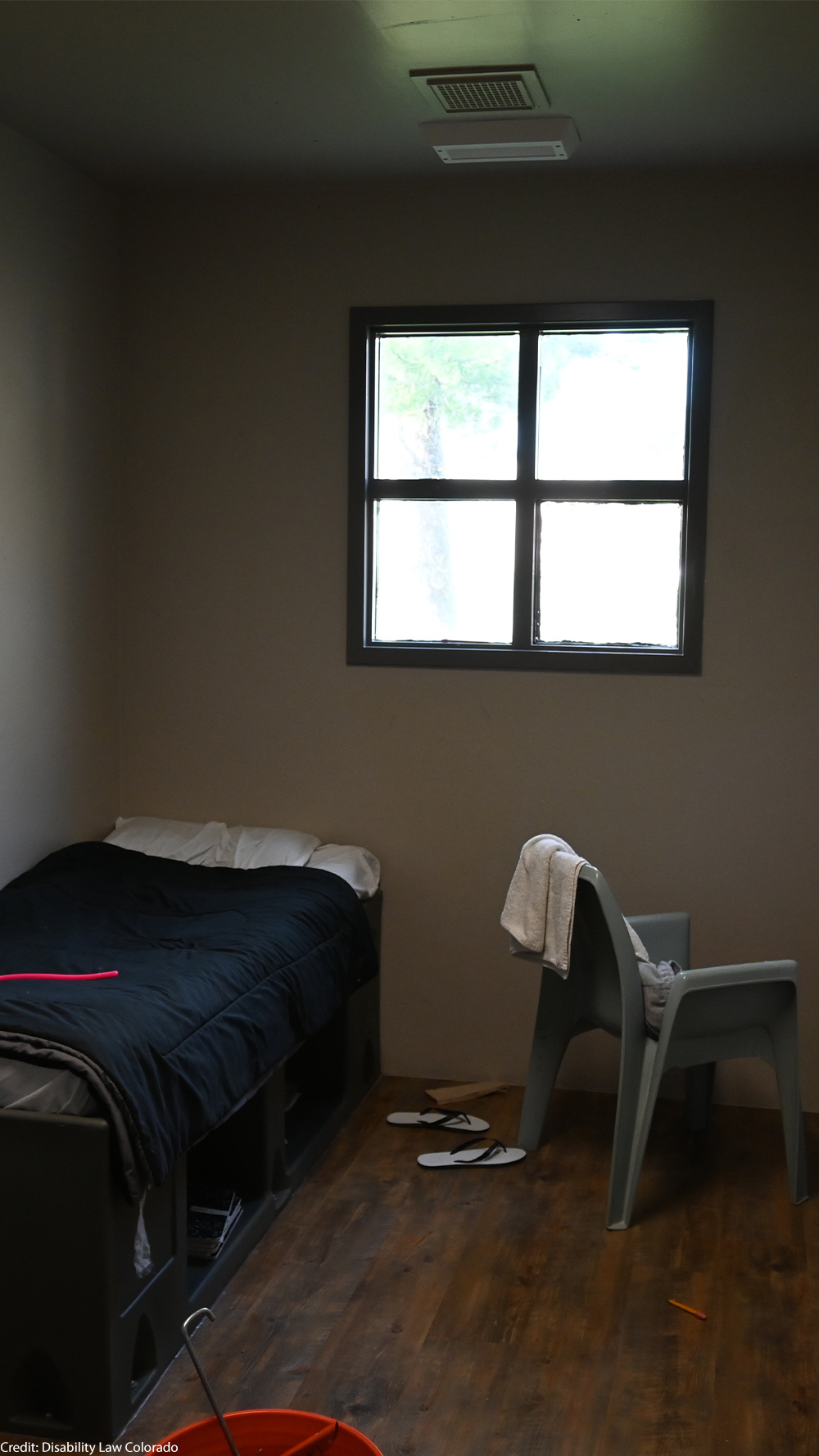 A small, sparsely furnished room with a single bed, a plastic chair with a towel draped over it, a barred window, and a pair of flip-flops on the wooden floor.