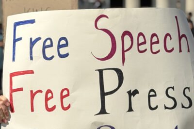 Person at a rally holds a large handmade sign reading “Free Speech, Free Press, Free People,” with a crowd and the Lincoln Memorial building in the background.