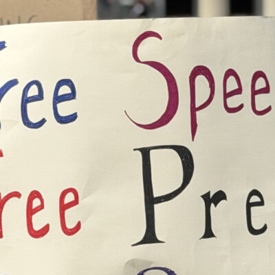Person at a rally holds a large handmade sign reading “Free Speech, Free Press, Free People,” with a crowd and the Lincoln Memorial building in the background.